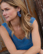 Woman wearing a blue dress with matching butterfly earrings, sitting against a wooden door.