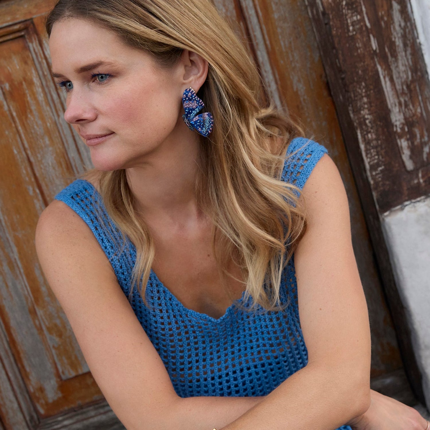 Woman wearing a blue dress with matching butterfly earrings, sitting against a wooden door.