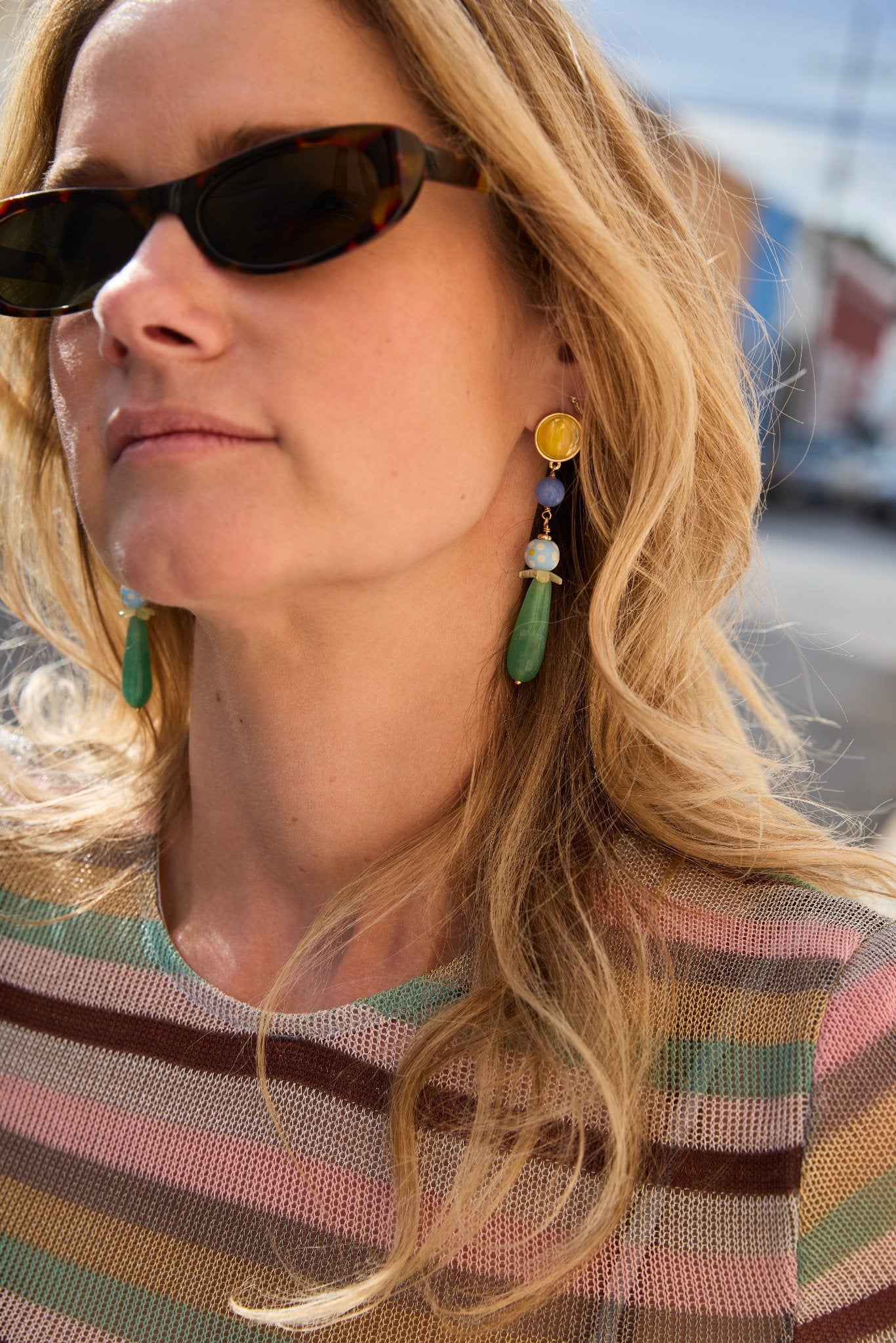Close-up of a person wearing sunglasses and colorful earrings with a blurred background