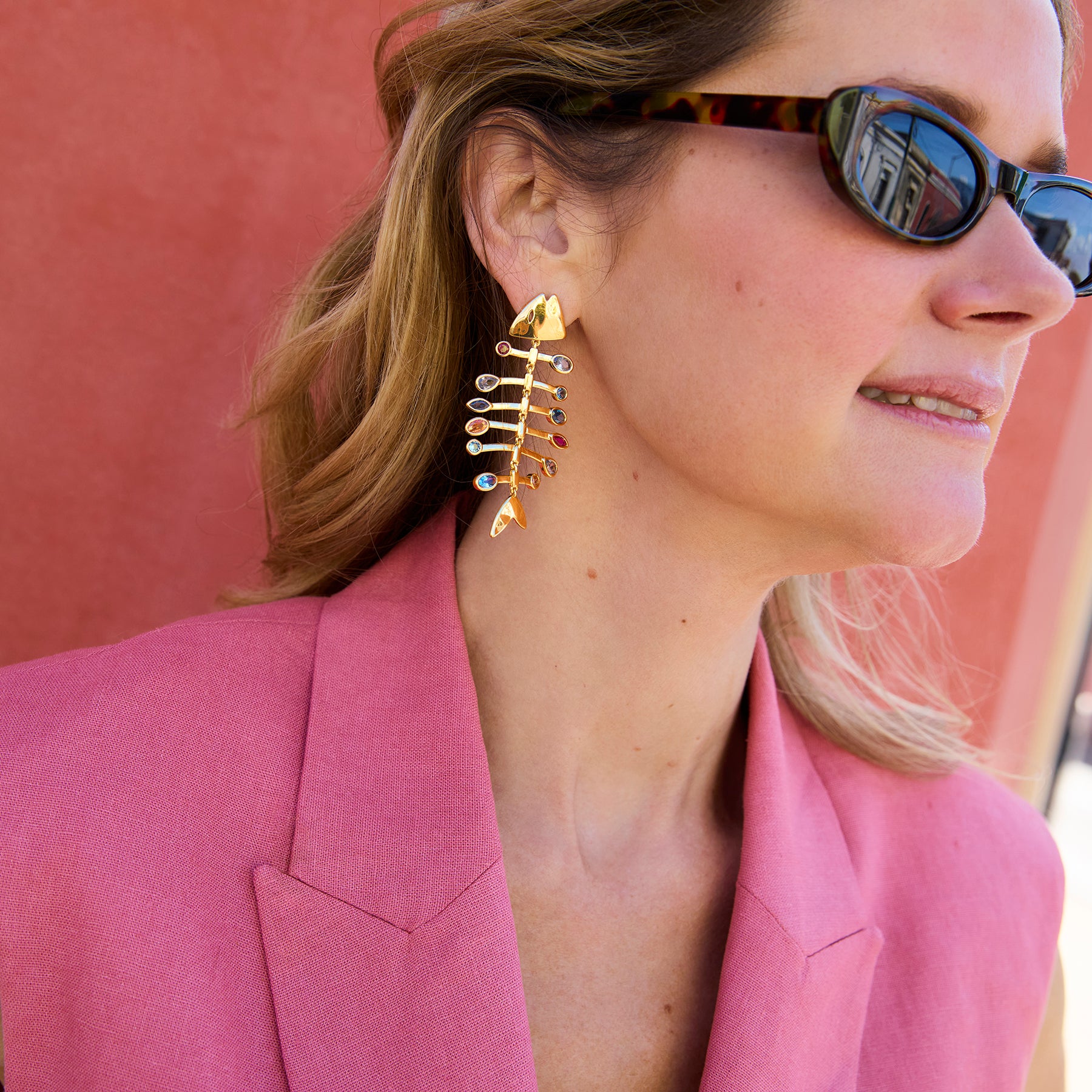 Woman wearing gold fishbone earrings and sunglasses with a pink background