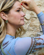 Woman with tattoos on her arm wearing a light blue top with a gold cuff bracelet on her wrist, against a stone wall background