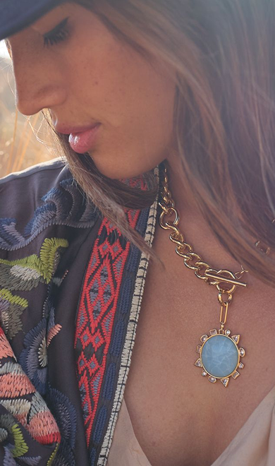 Close-up of a person wearing a gold necklace with a blue pendant, set against a blurred natural background.