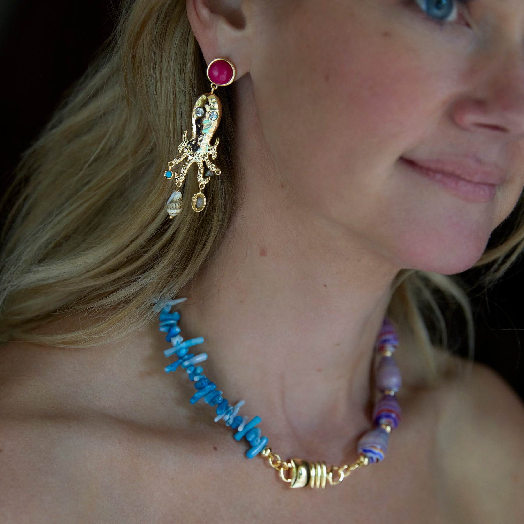 Close-up of a woman wearing a blue beaded necklace and gold earrings with a red gemstone.