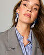 A close-up of a woman wearing large, dangling earrings with blue beaded tops and white tassel fringe, styled with a gray blazer and a blue-and-white striped button-down shirt.