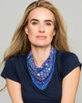 A woman with blonde hair wearing a navy blue t-shirt and a blue patterned neck scarf with colorful geometric designs and blue pom-pom trim, set against a solid light gray background.