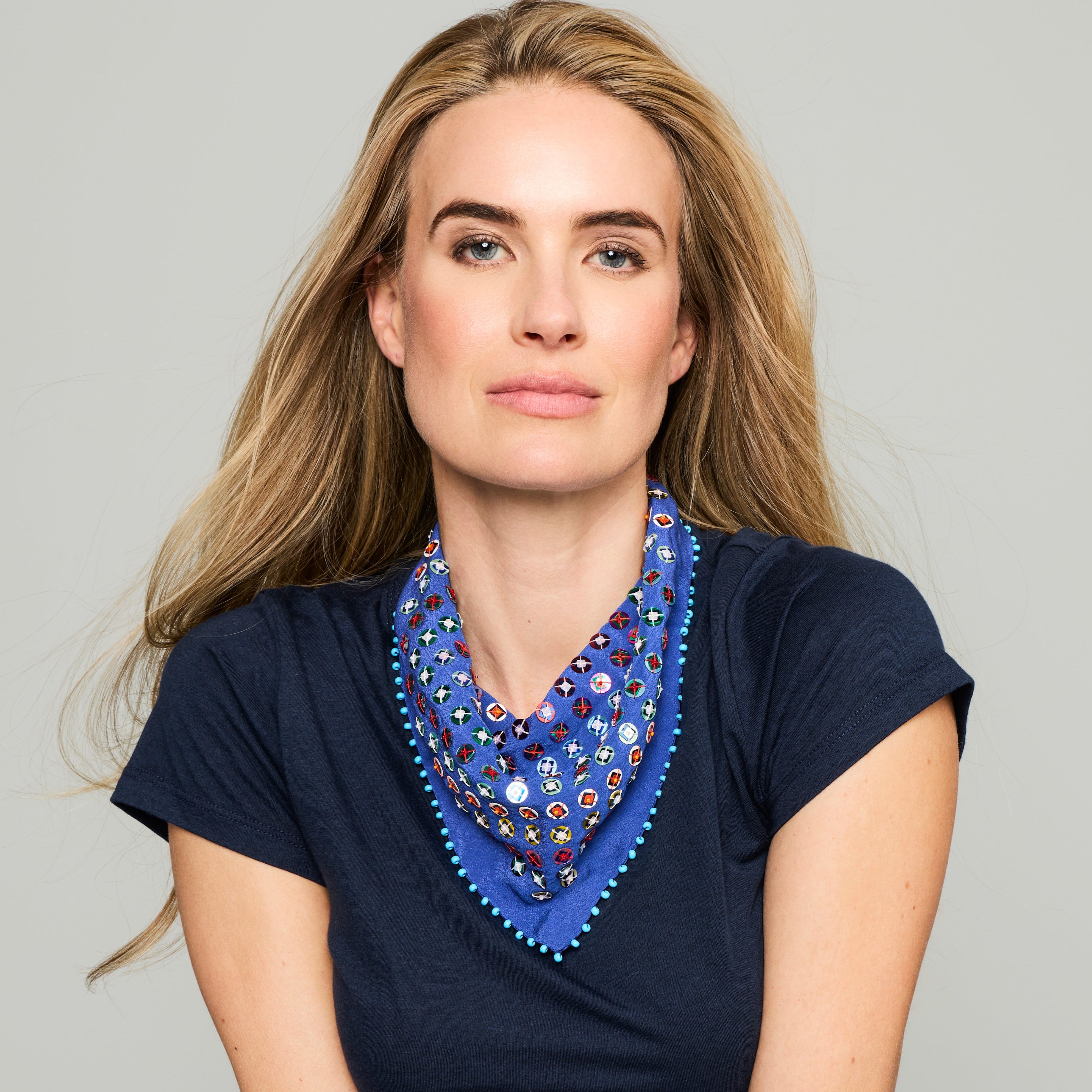 A woman with blonde hair wearing a navy blue t-shirt and a blue patterned neck scarf with colorful geometric designs and blue pom-pom trim, set against a solid light gray background.