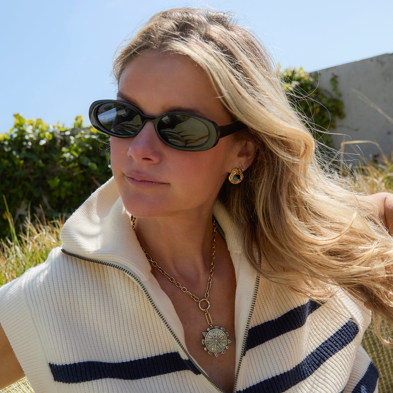 Woman wearing sunglasses and gold stud earrings with a white sweater with blue stripes outdoors
