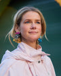 Woman in a light jacket standing in front of a yellow wall with an archway wearing fish shaped embroidered earrings