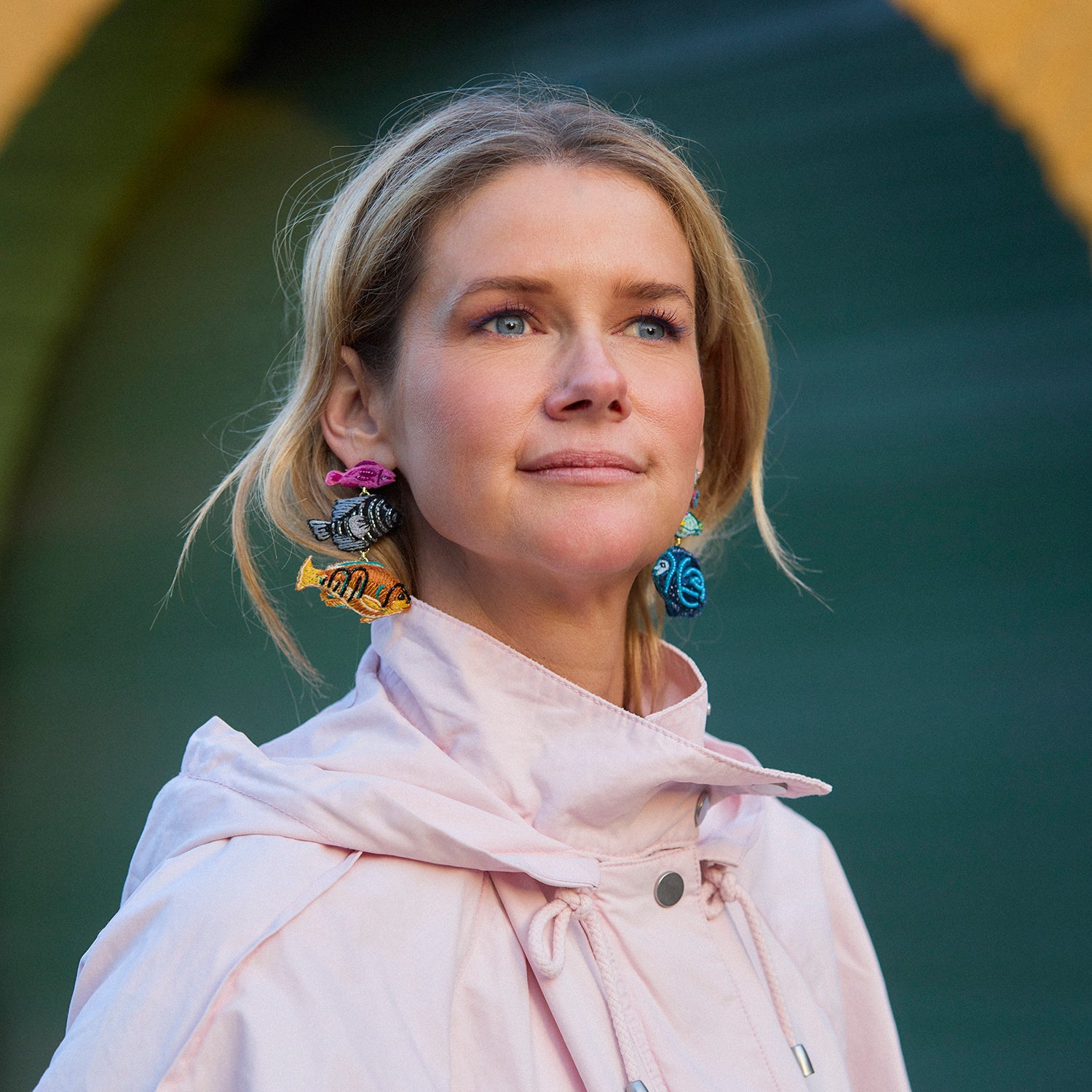 Woman in a light jacket standing in front of a yellow wall with an archway wearing fish shaped embroidered earrings