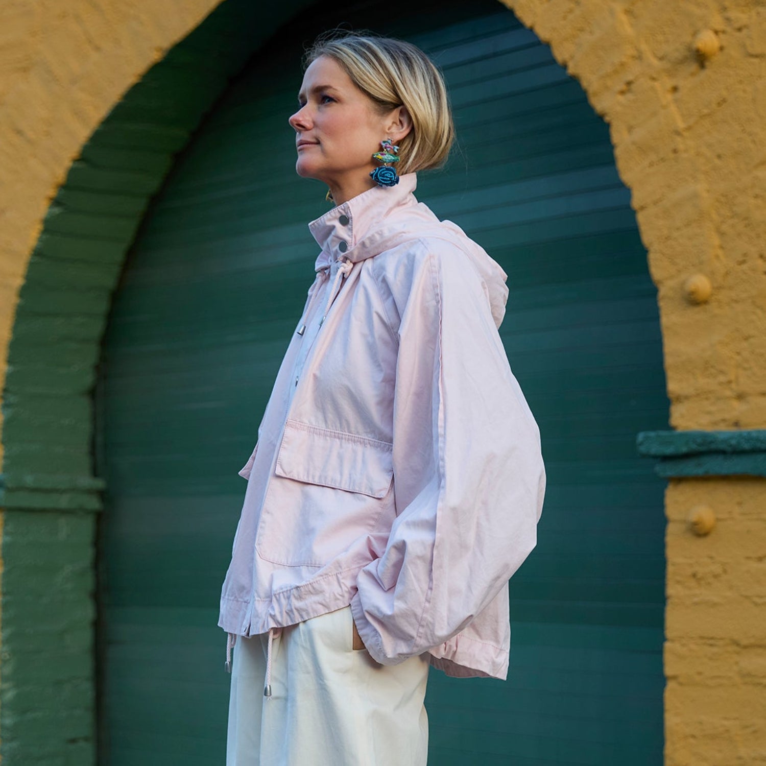 Woman in a light jacket standing in front of a yellow wall with an archway wearing fish shaped embroidered earrings