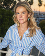 Woman in blue shirt with wearing gold and charm earrings against palm tree background