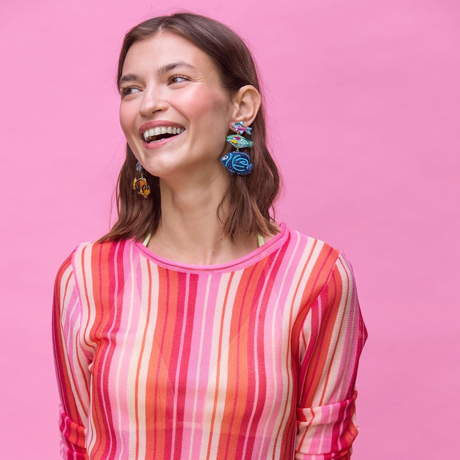 Woman wearing a colorful striped shirt with fish shaped colorful beaded earrings against a pink background