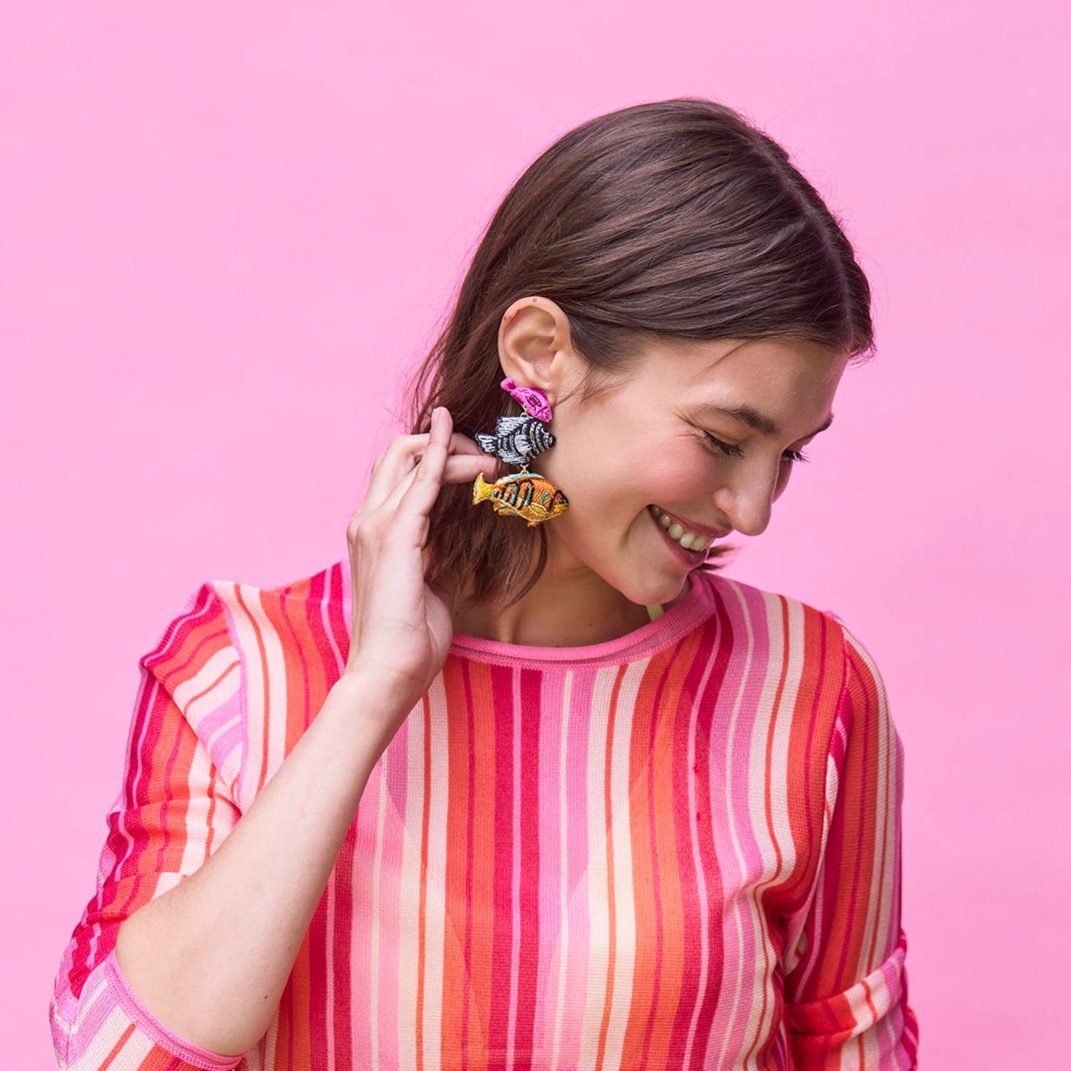 Woman wearing a colorful striped shirt with fish shaped colorful beaded earrings against a pink background