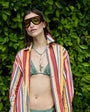 Woman wearing a green bikini top with a colorful striped shirt over it, wearing a beaded necklace standing against a green leafy background.