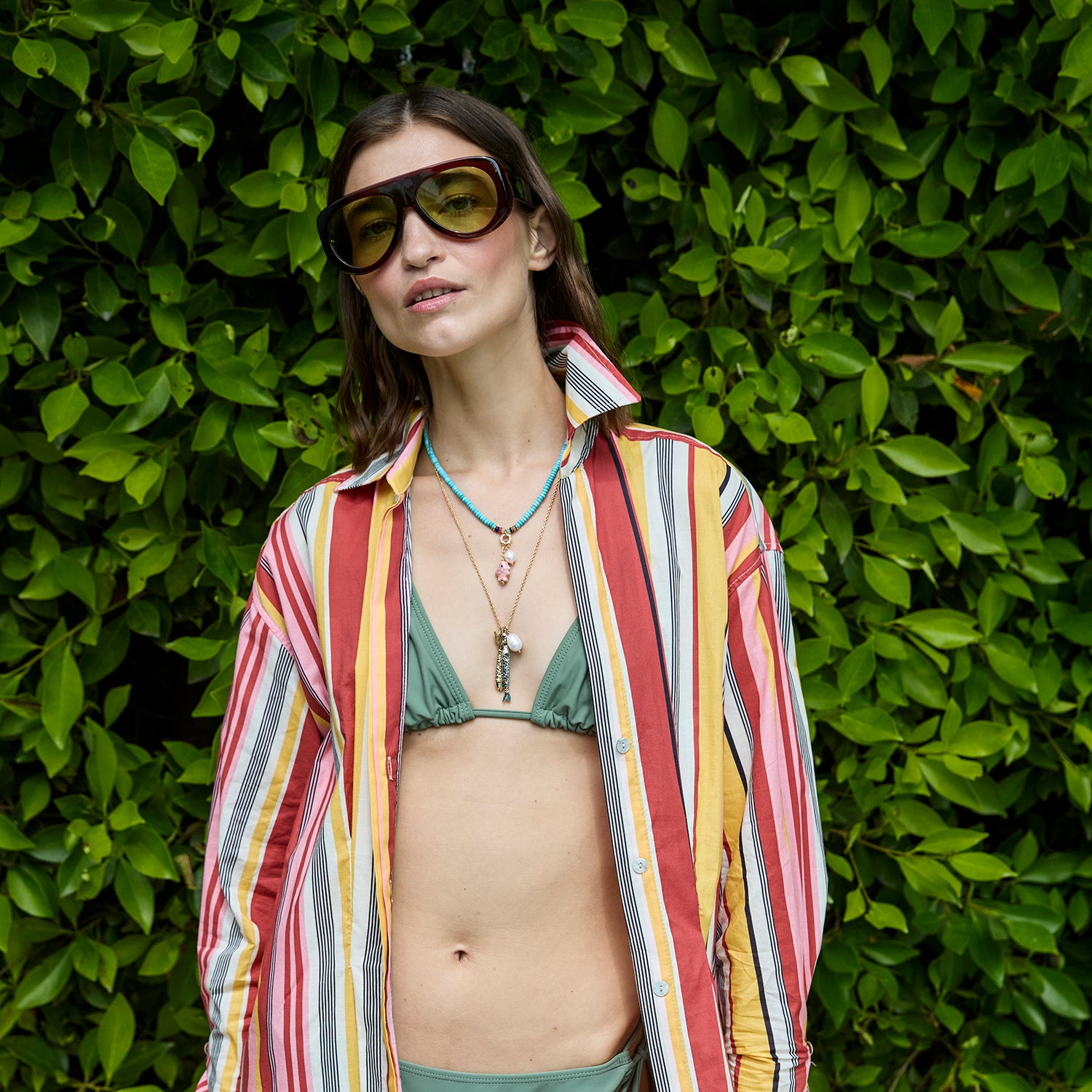 Woman wearing a green bikini top with a colorful striped shirt over it, wearing a beaded necklace standing against a green leafy background.