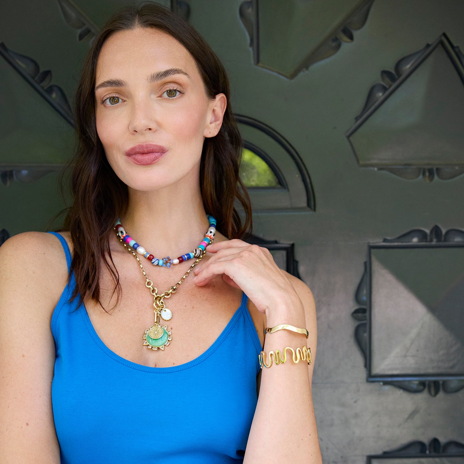 Woman wearing colorful and beaded jewelry against a decorative wall.
