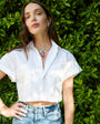 Woman wearing a white shirt and blue jeans with a gold collar necklace featuring fish shaped charms standing against a green hedge.