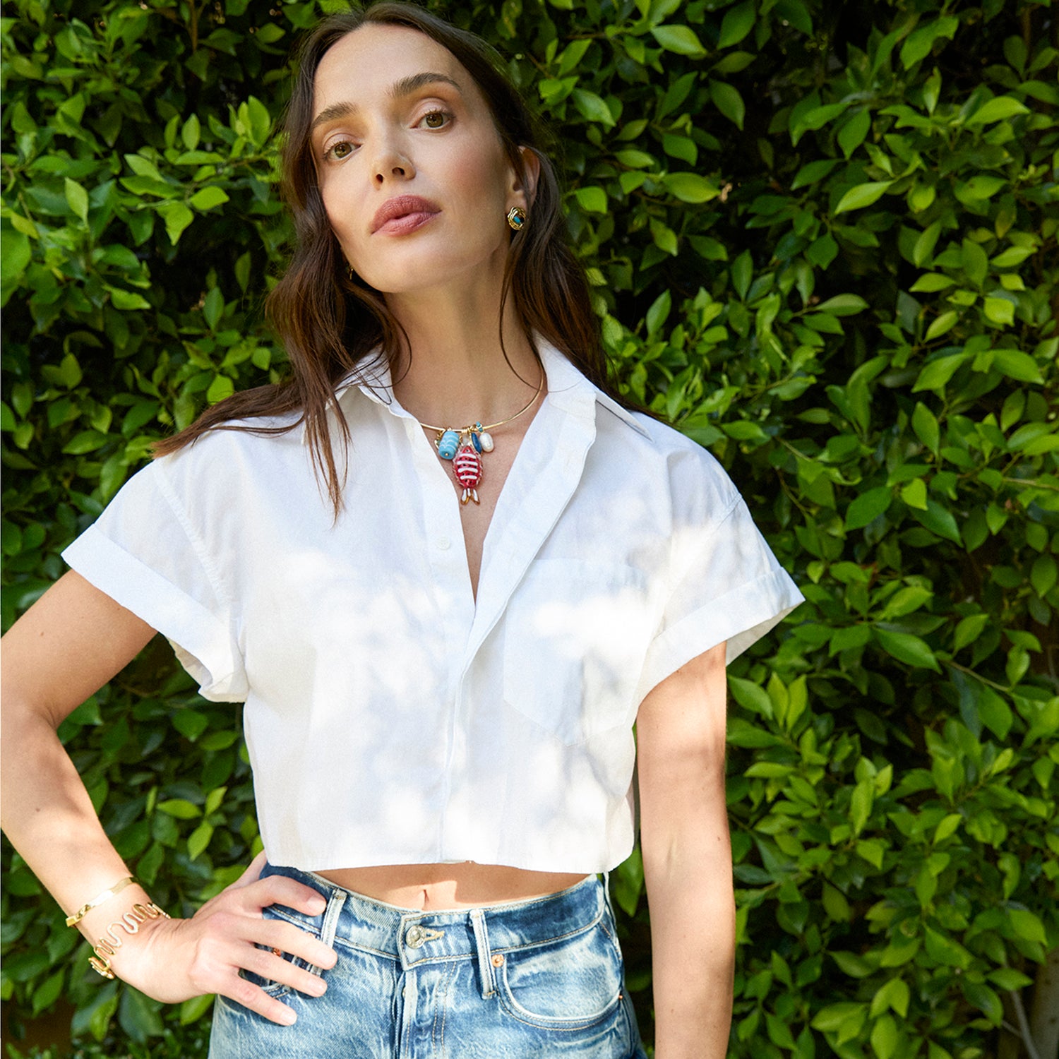 Woman wearing a white shirt and blue jeans with a gold collar necklace featuring fish shaped charms standing against a green hedge.