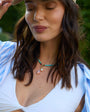 Woman wearing a white top and blue striped shirt with a beaded necklace, outdoors.