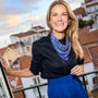 A smiling woman with blonde hair stands on a balcony wearing a navy blue blouse, a bright blue skirt, and a patterned blue scarf with white and yellow details. The background shows a scenic view of white buildings with red terracotta roofs under a cloudy sky.