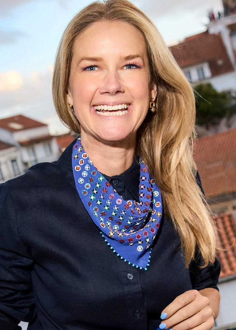 A smiling woman with blonde hair stands on a balcony wearing a navy blue blouse, a bright blue skirt, and a patterned blue neck scarf with white and yellow details. The background shows a scenic view of white buildings with red terracotta roofs under a cloudy sky.