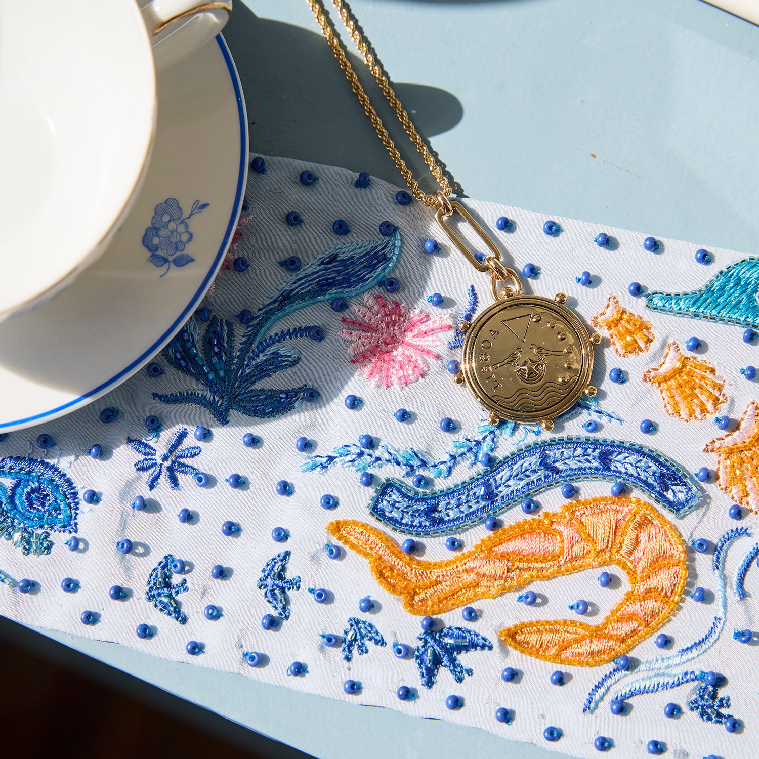 A gold-toned necklace with a round medallion pendant is laid on a white embroidered textile featuring blue and orange sea creature embroidery, next to a white teacup and saucer on a pale blue surface.