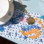 A gold-toned necklace with a round medallion pendant is laid on a white embroidered textile featuring blue and orange sea creature embroidery, next to a white teacup and saucer on a pale blue surface.