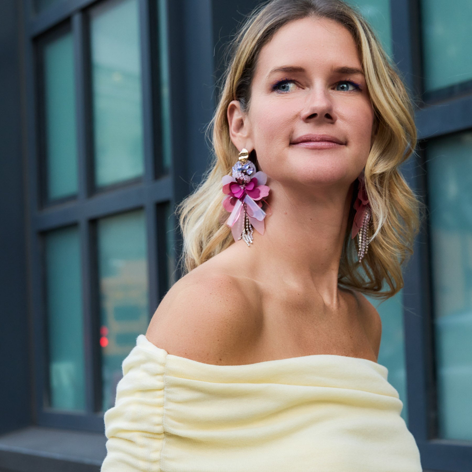 Maggie wearing a yellow off-shoulder top with floral earrings in front of a building.
