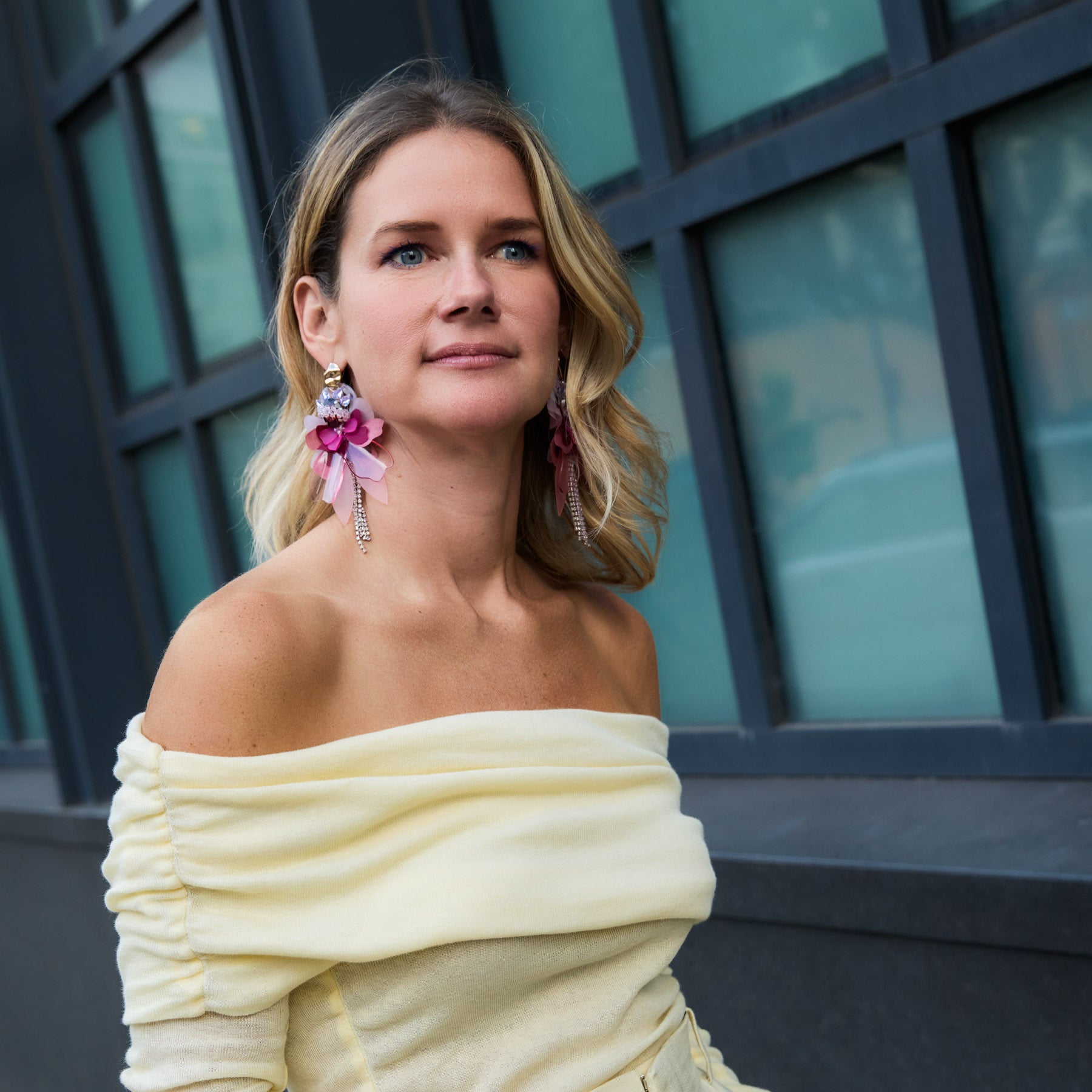 Maggie wearing a yellow off-shoulder top with floral earrings in front of a building.
