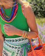 Woman in a green top with colorful necklace and patterned skirt, holding an orange bag, standing in a garden.