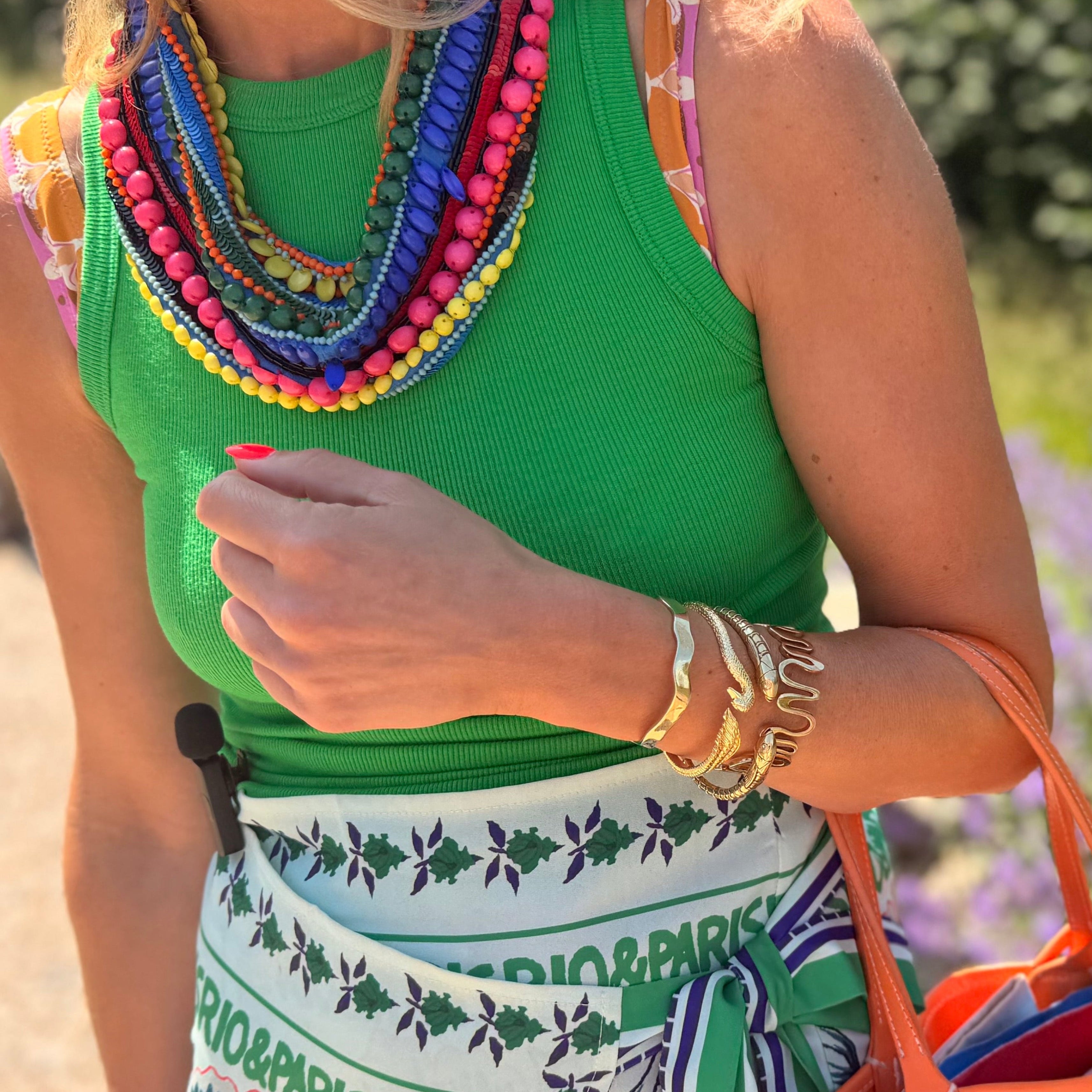 Woman in a green top with colorful necklace and patterned skirt, holding an orange bag, standing in a garden.