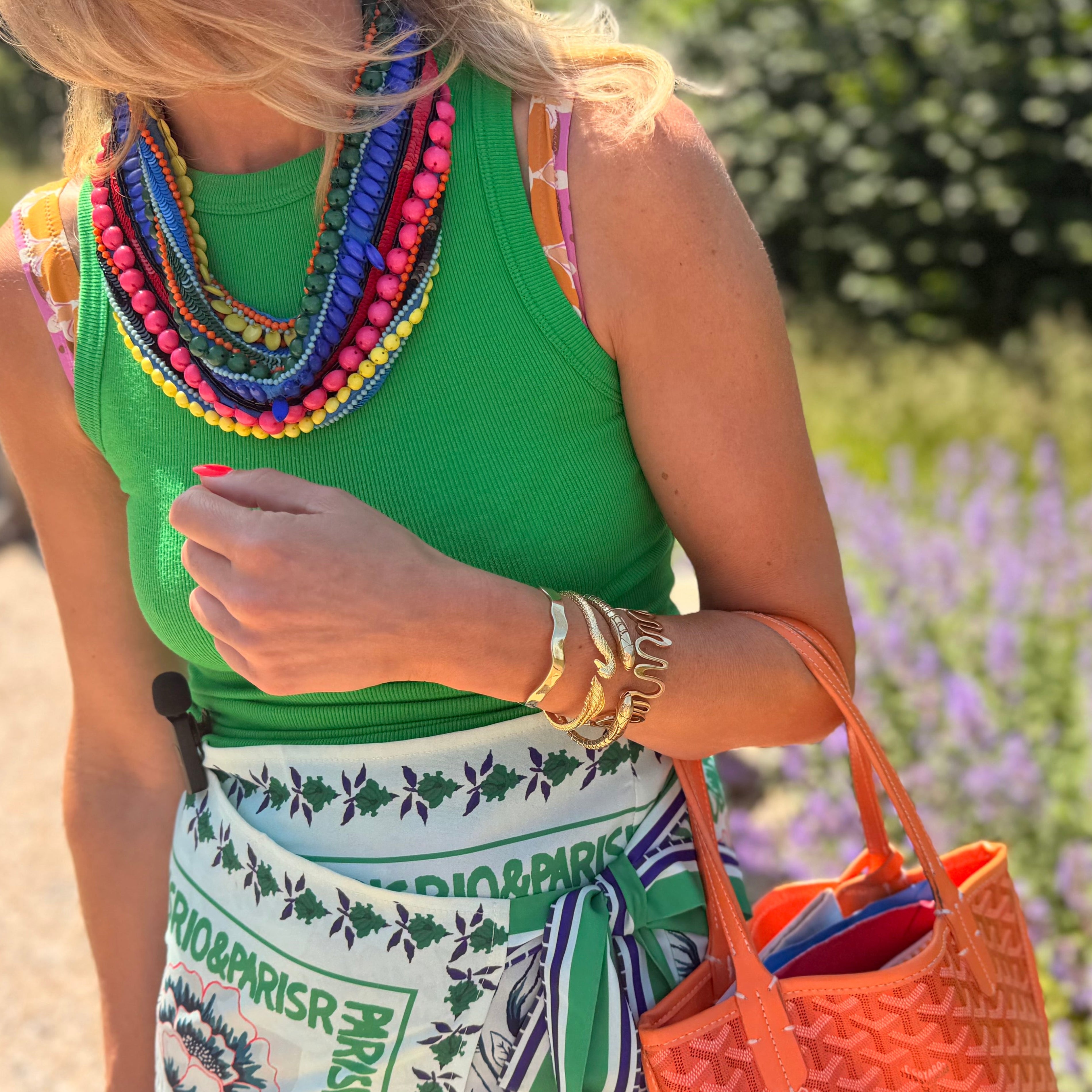 Woman in a green top with colorful necklace and patterned skirt, holding an orange bag, standing in a garden.