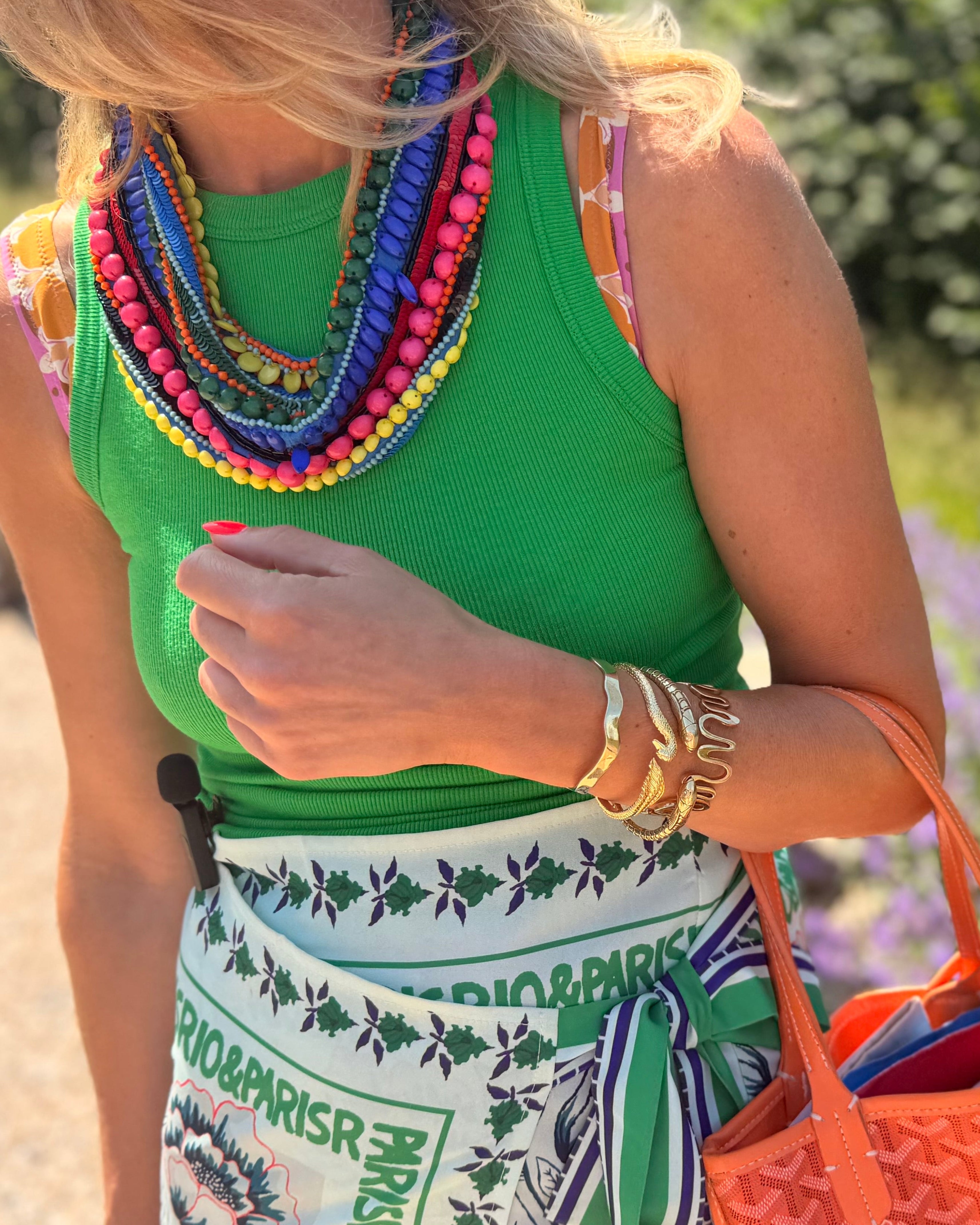 Woman in a green top with colorful necklace and patterned skirt, holding an orange bag, standing in a garden.