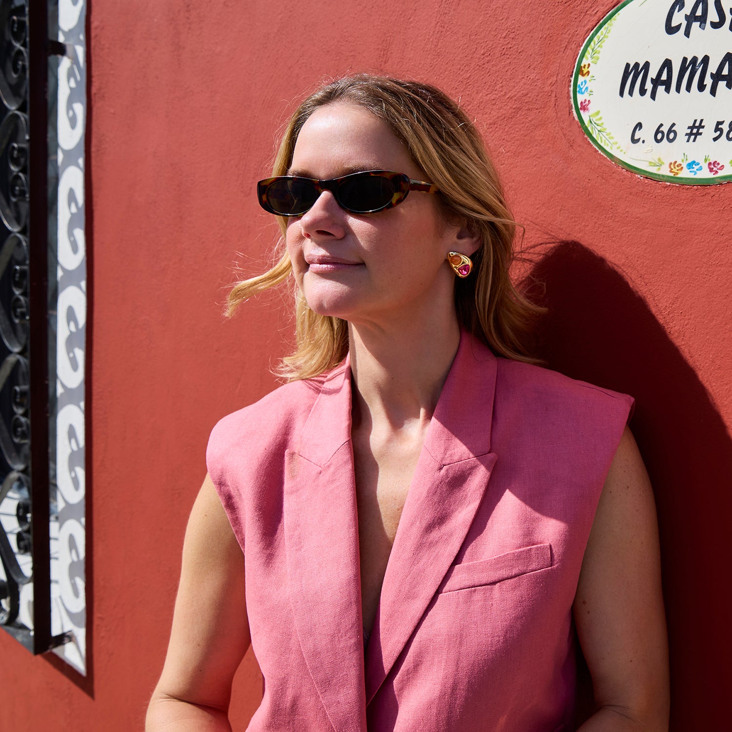 Woman in a pink sleeveless blazer standing against a red wall with a sign wearing gold and multicolor stud earrings