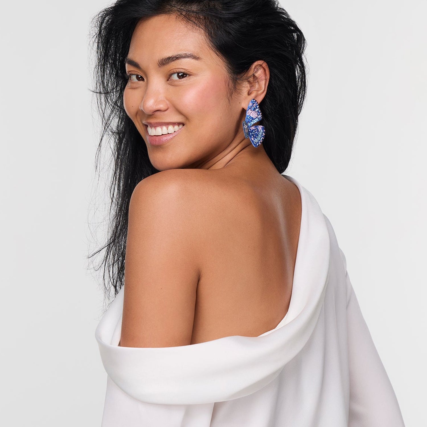 Woman with butterfly earring in ear while wearing a  white garment draped over her shoulders against a plain background