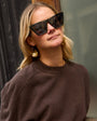 Woman wearing sunglasses and a brown sweater with gold sequin hoop earrings against a blurred background