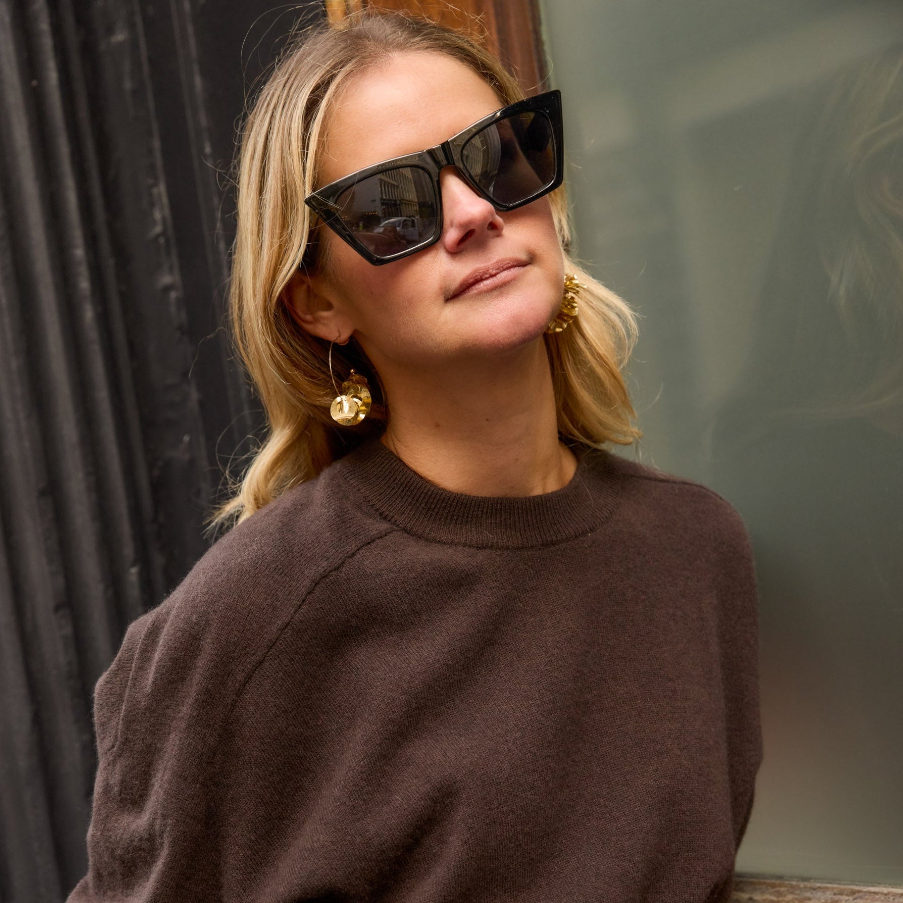 Woman wearing sunglasses and a brown sweater with gold sequin hoop earrings against a blurred background