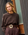 Woman in a brown outfit and gold sequin hoop earrings standing against a textured wall.