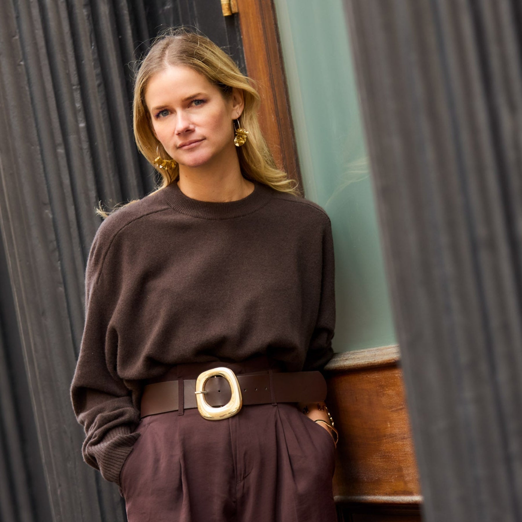 Woman in a brown outfit and gold sequin hoop earrings standing against a textured wall.