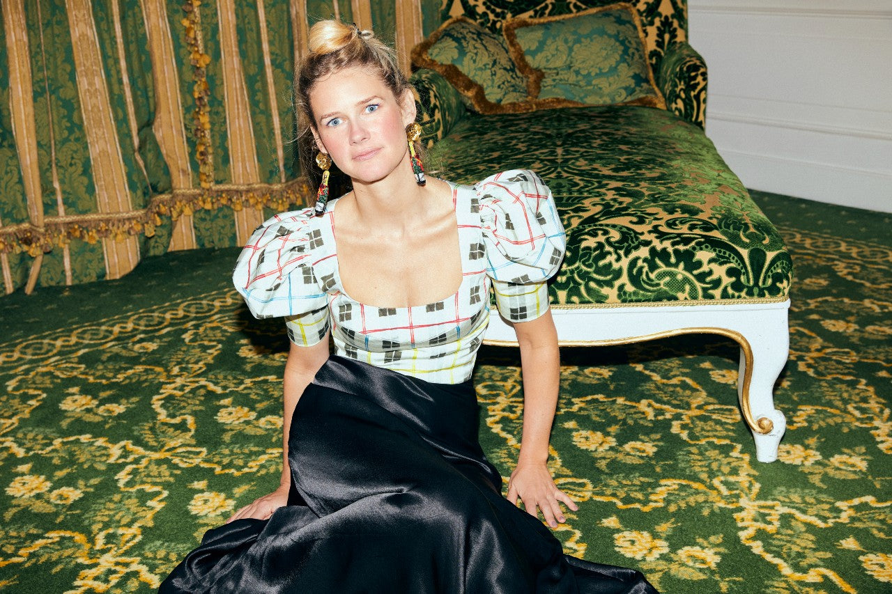 Maggie in a patterned top, black skirt, and champagne bottle earrings, sitting on a ornate green sofa.