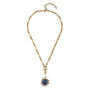 A gold chain necklace with a central pendant featuring a round blue stone surrounded by small clear stones, displayed on a clean white background.