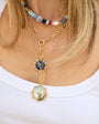 A close-up of a woman wearing a layered gold necklace set. The set includes a short beaded choker, a longer chain with a blue stone pendant, and a gold pocket-watch pendant necklace, all worn over a white tank top.
