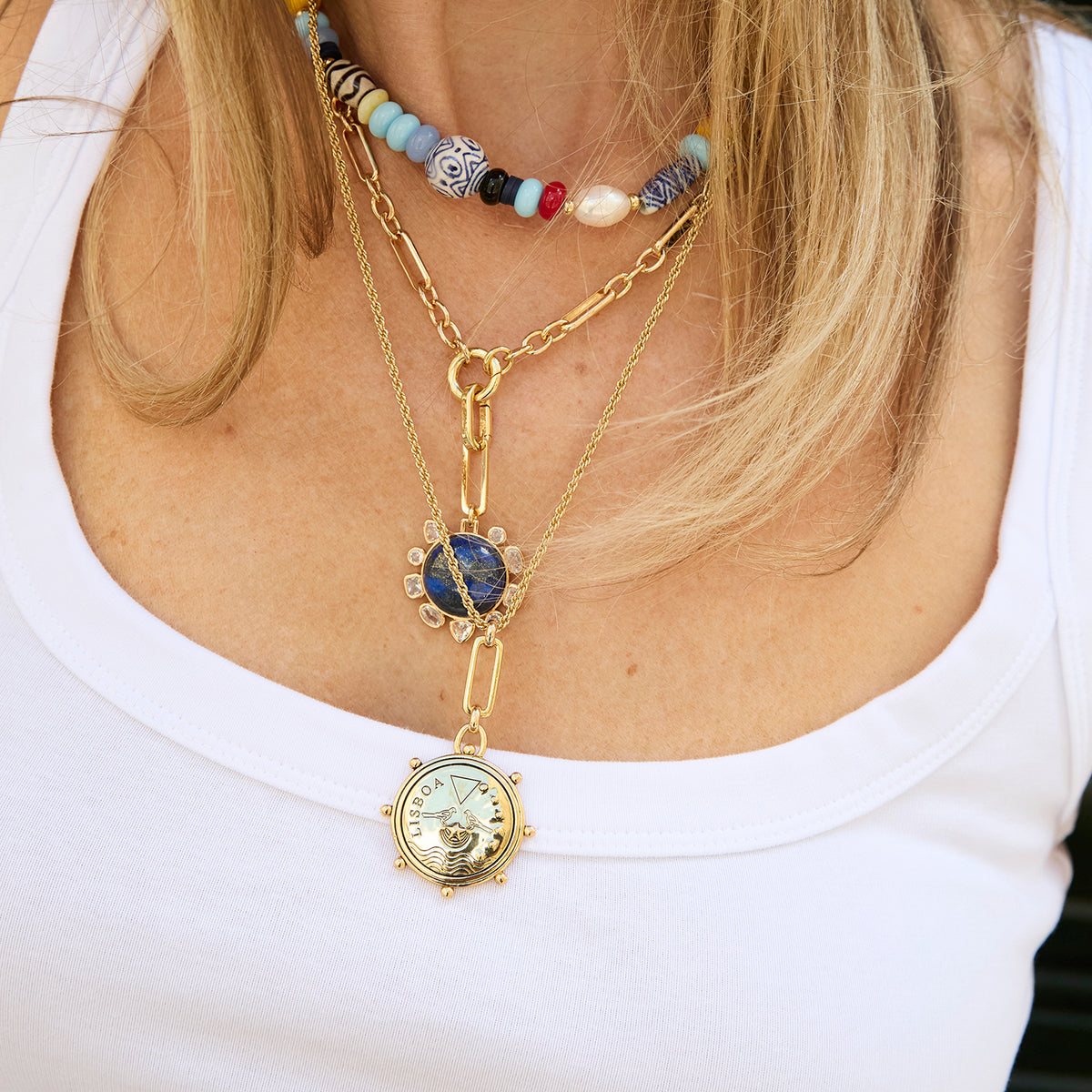 A close-up of a woman wearing a layered gold necklace set. The set includes a short beaded choker, a longer chain with a blue stone pendant, and a gold pocket-watch pendant necklace, all worn over a white tank top.