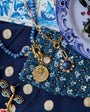 A styled flat-lay of jewelry and accessories, featuring a gold necklace with celestial pendants, blue beaded necklaces, gold and blue earrings, and a blue patterned textile on a white and blue surface.