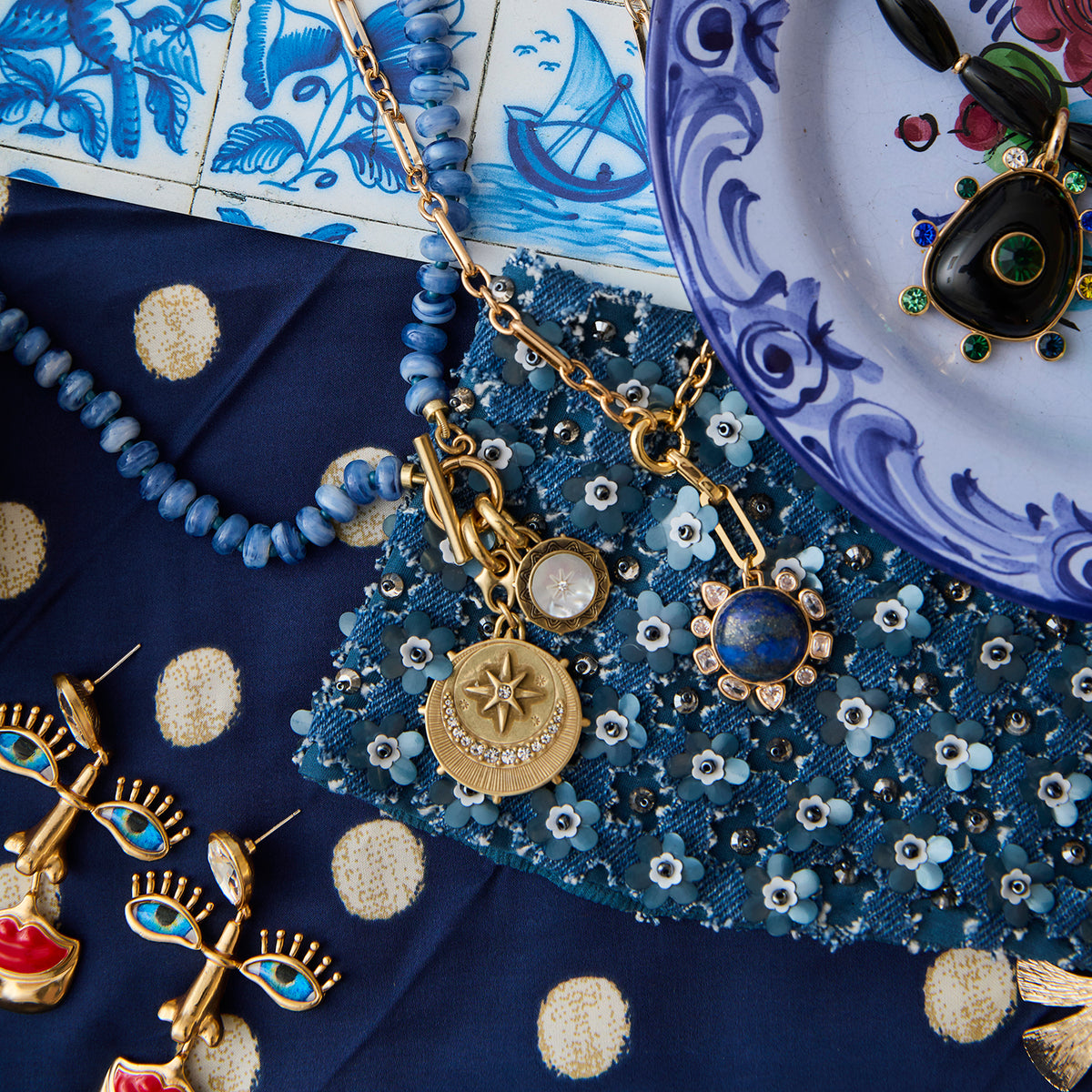 A styled flat-lay of jewelry and accessories, featuring a gold necklace with celestial pendants, blue beaded necklaces, gold and blue earrings, and a blue patterned textile on a white and blue surface.