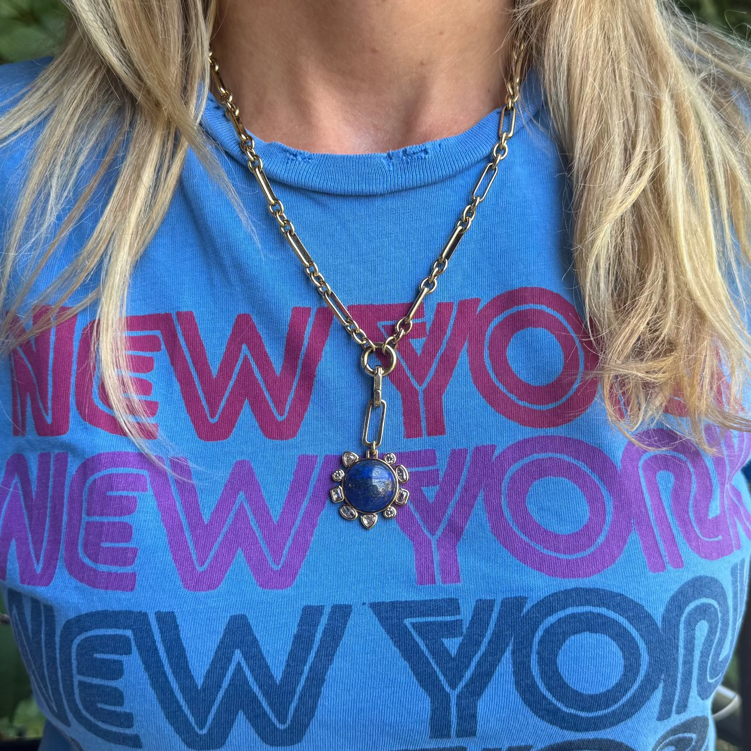 A close-up of a person wearing a bright blue graphic t-shirt with stacked 'NEW YORK' text in pink, purple, and navy, accessorized with a gold chain necklace featuring a blue sun pendant.