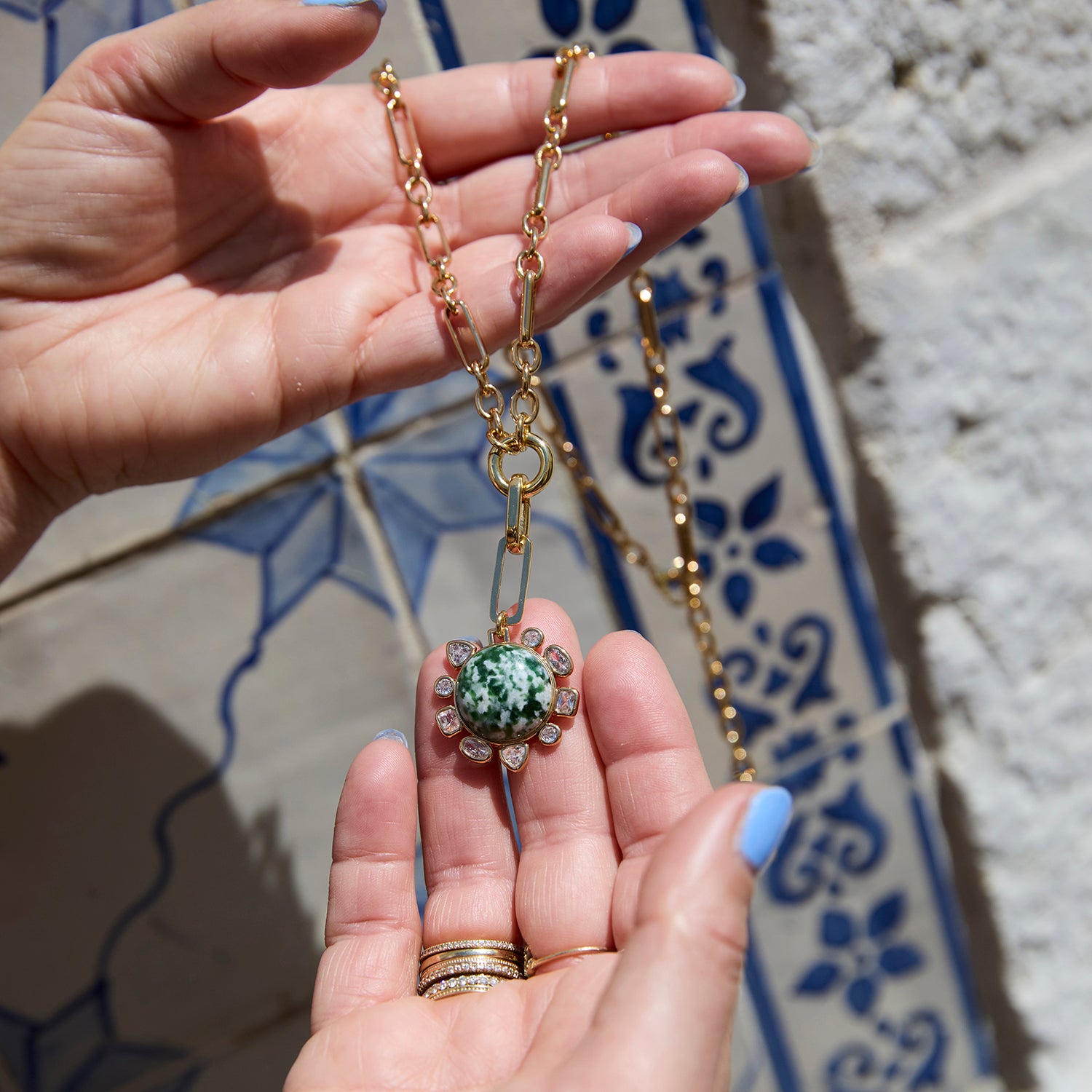 A gold chain necklace with a central green marbled stone pendant and small clear stones, held in a person's hand against a blue and white tiled background.