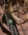 A close-up shot of a gold-tone pendant necklace with a green and white marbled cabochon stone, styled on a person's chest over a brown polka-dot jacket and a black-and-brown checkered shirt.