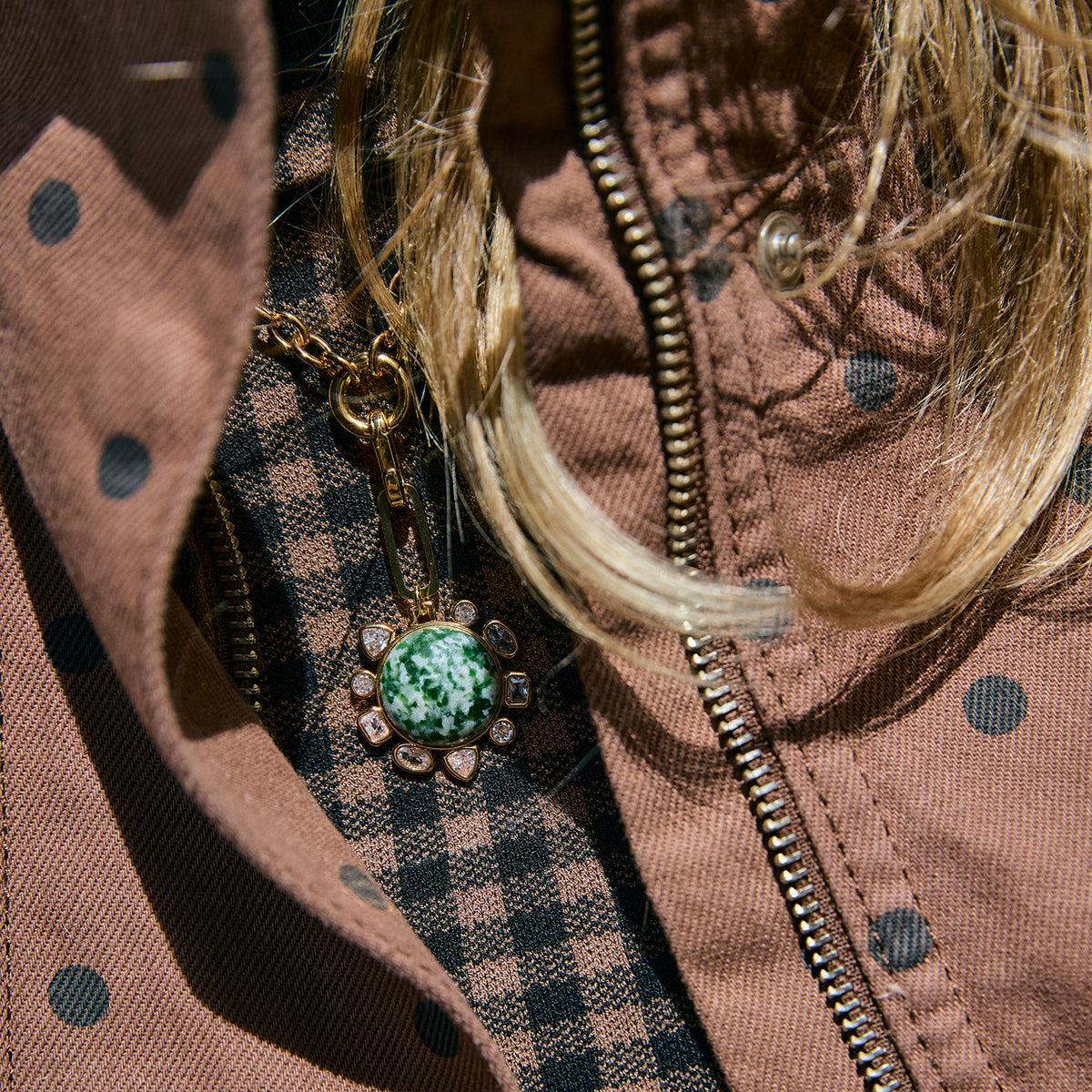 A close-up shot of a gold-tone pendant necklace with a green and white marbled cabochon stone, styled on a person's chest over a brown polka-dot jacket and a black-and-brown checkered shirt.