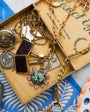 A vintage-style jewelry box filled with antique gold-toned necklaces, pendants, and charms, including a green stone pendant and coin-like charms, set against a blue patterned textile background.